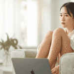 Young woman sitting with a laptop on her thighs, her face clearly visible, showing early signs of Toasted Skin Syndrome with a light net-like reddish-brown pattern on her legs.